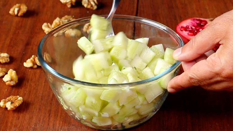 A spoon is seen tossing cucumbers with salad dressing in a large glass bowl while a hand holds the bowl steady on a wooden counter. California walnut kernels and an open pomegranate lie in the background - Konkani Cucumber and Pomegranate Salad Recipe